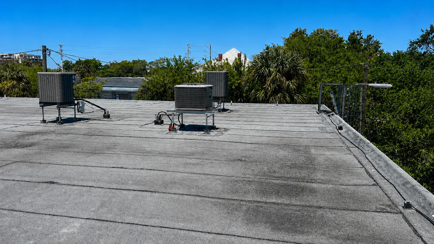 Wide-angle photograph of a commercial flat roof in Tampa Bay featuring multiple rooftop HVAC units installed on support stands. The light gray roofing membrane shows visible weathering, dark moisture-related staining, surface discoloration, and areas of wear surrounding the mechanical equipment. Electrical conduits and service lines run across the roof surface, while parapet edge flashing borders the perimeter. In the background, mature trees, utility poles, overhead power lines, and neighboring buildings are visible beneath a clear blue sky. The image illustrates common signs of aging and potential maintenance concerns on a commercial roofing system, including membrane deterioration, staining around rooftop equipment, and conditions that may indicate the need for professional commercial roof repair and inspection by a qualified roofing contractor in Tampa Bay.