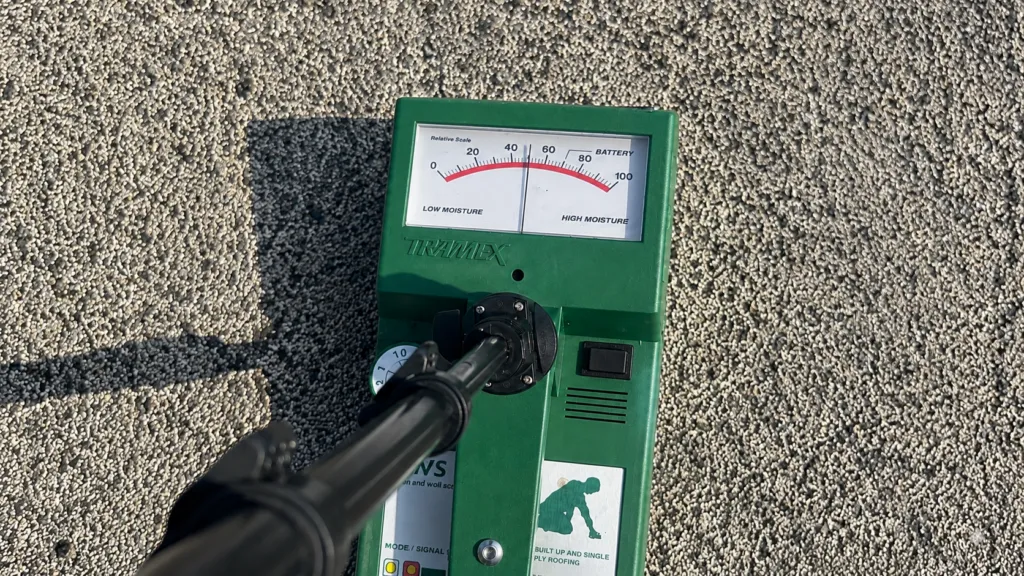 Close-up photograph of a green Tramex moisture meter positioned on a light gray granulated roofing surface during a roof inspection. The handheld instrument features an analog gauge labeled with a relative moisture scale ranging from 0 to 100, with the needle positioned near the mid-range reading. A black extension handle is attached to the center of the device and extends diagonally toward the foreground, partially obscuring the lower portion of the meter. Strong sunlight casts a defined shadow of the handle and instrument across the textured roof surface, highlighting the granular roofing aggregate. The meter housing includes control markings, indicator labels, and a graphic showing a worker kneeling. The image emphasizes moisture detection equipment in use on a commercial low-slope roofing system, with detailed visibility of the roofing texture, instrument components, and inspection conditions.